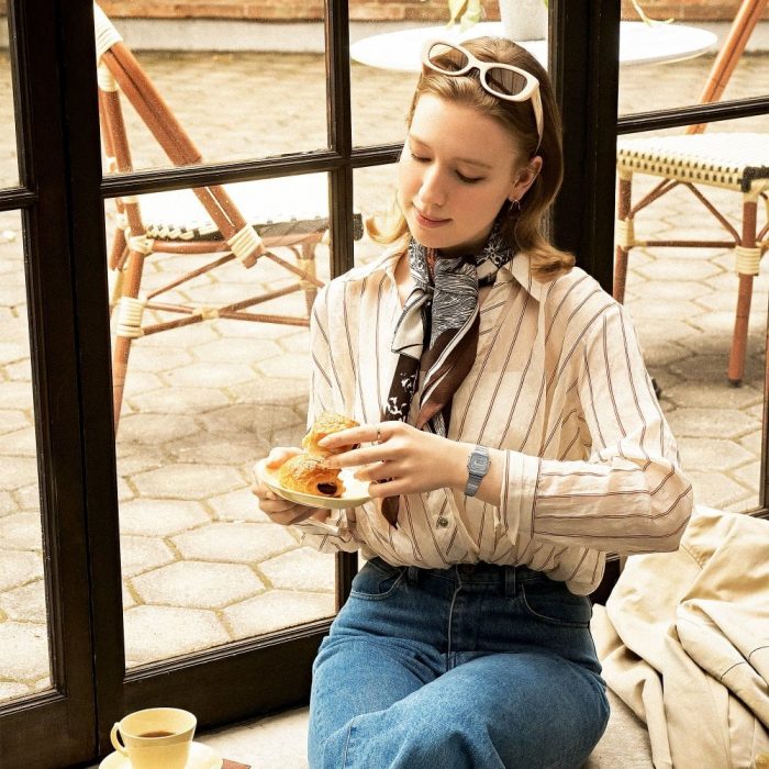 Woman wearing the Casio Ladies Vintage Mini (LA670) with a vintage style sunglasses, shirt, jeans and a neck scarf eating a croissant.