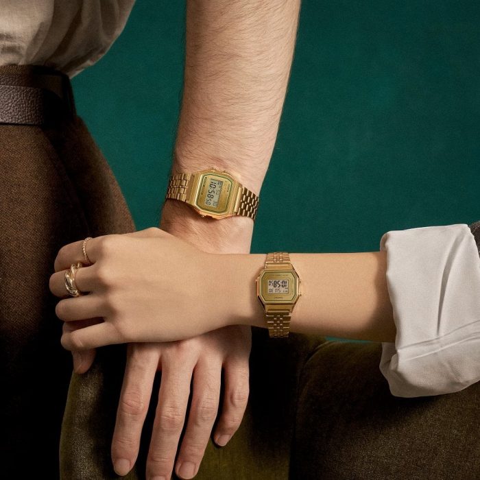 A women and a man wearing the Casio gold LA680 with green woolen trousers a smart tucked in shirt and a cuffed blazer and matching gold jewellery.